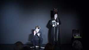 Szene aus Leonce und Lena. Prince Leonce sitz mit einem goldenen Lorbeerkranz auf einem Hocker. Neben ihm steht die Hofadame mit einem Stapel Bücher in der Hand.