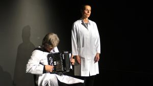 Ein Mann sitzt und rechts von ihm steht eine Frau. Beide tragen einen weißen Laborkittel. Es sind zwei Musiker, die auf der Bühnen Labormusik spielen. Er spielt Ziehharmonika und sie singt.