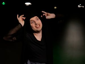 Ein Schauspieler auf schwarzer Bühne schaut mit staunendem Gesicht nach rechts. Er trägt eine schwarze Kappe. Sein Kostüm ist ebenfalls schwarz.