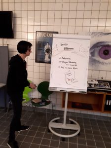 Susanne am Flipchart im Casino des TheaterLabor TraumGesicht. Auf dem Bogen ist zu lesen: Herzlich Willkommen. Ankommen, Dragon Dreaming und auf zu neuen Ufern.