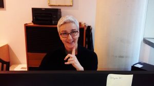 Antje, eine Frau sitzt vor einem Büroschrank mit schwarzem Rollo. Darauf steht ein Drucker. Sie trägt eine Brille und lacht in die Kamera.