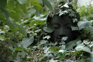 Buddhakop in einem Staudengarten. Umwuchert von Efeu.