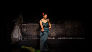 Eine junge Frau tanzt auf der Bühne. Sie trägt eine rote Blume in der Hochsteckfrisur und trägt ein grünes Abendkleid mit Pailletten.