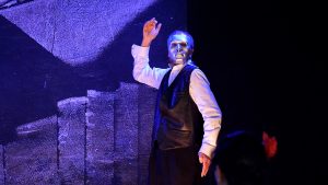 Ein Schauspieler mit einer silbernen Maske steht auf der bläulich beleuchteten Bühne. Er zeigt eine starke Gebärde: die rechte Hand schwebt über seinem Kopf und die linke deutet in den Boden.
