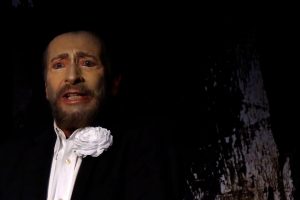 Ein Schauspieler mit stark geschminktem Gesicht zeigt einen betroffenen Ausdruck. Er trägt ein weißes Hemd eine dunkle Jacke und eine weiße Blume im Revier Wolfgang Keuter als Tod in Der Tor und der Tod.