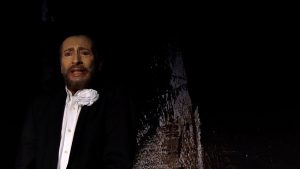 Ein Schauspieler mit stark geschminktem Gesicht zeigt einen betroffenen Ausdruck. Er trägt ein weißes Hemd eine dunkle Jacke und eine weiße Blume im Revier Wolfgang Keuter als Tod in Der Tor und der Tod.