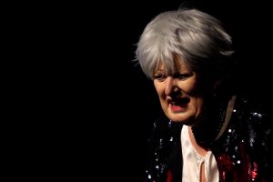 Eine Schauspielerin steht im schwarzen Bühnenraum. Sie trägt eine rot-schwarze Pailletten-Jacke. Sie hat weiße Haare in einem modernem Schnitt und schaut mit auf den Boden geneigtem Blick nachdenklich in die Welt. Doris Horn als Mutter in Der Tor und der Tod.