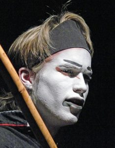 Florian Frowein als Hauptmann Fukakusa. Ein junger Mann mit blonden Haaren und verfremdeter Schminkmaske nach asiatischer Vorlage schaut ernst in den dunklen Bühnenraum. Er schultert eine Bambusstange.