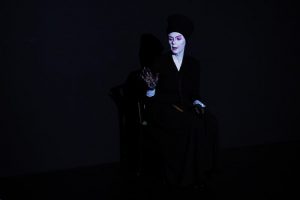 Im schwarzen Bühnenraum sitzt eine weiß geschminkte Frau und schaut nach links mit einer erhobenen Hand