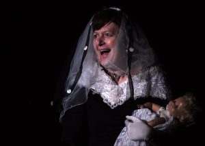 Im schwarzem Bühnenraum. Eine Frau schaut lachend nach links mit einer Puppe in der Hand. Die Frau trägt einen weißen und schwaren Schleier, die Puppe ein weißes Taufgewand