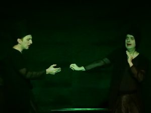 Zwie in schwarz gekleidete Figuren geben sich die Hand und lösen gerade den Handschlag. Sie stehen auf der schwarzen Bühne und sind zum Teil grün beleuchtet.