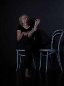 Eine Frau sitzt lachend auf einem grauen Stuhl. Sie zeigt ihre rechte Fusssohle. Sie trägt schwarze Übungskleidung und befindet sich auf der Bühne.