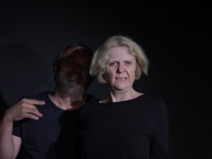 Ein Mann steht hinter einer Frau. Sie zeitgt ein entsetztes Gesicht. Beide tragen schwarze Übungskleidung und sind im schwarzem Bühnenraum.
