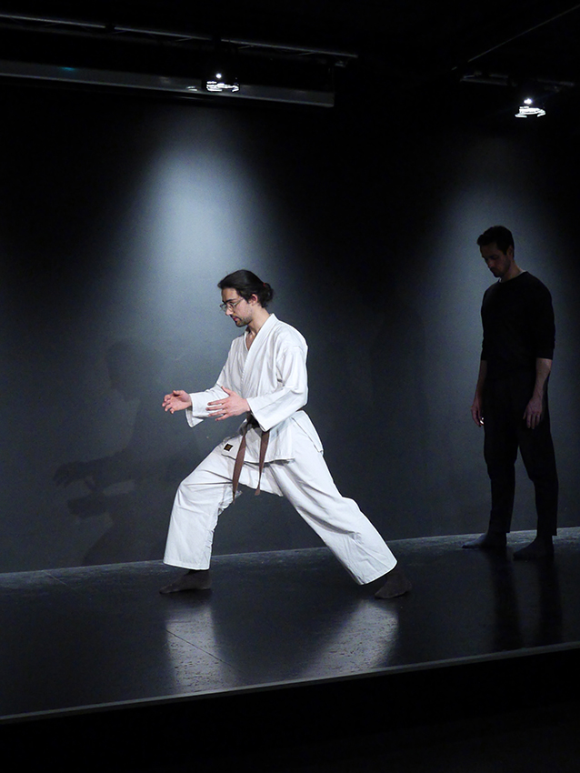Vorne links steht ein junger Lehrer und zeigt einen Karatestand. Im Hintergrund steht ein Mann in schwarzer Übungskleidung und sieht ihm aufmerksam zu.