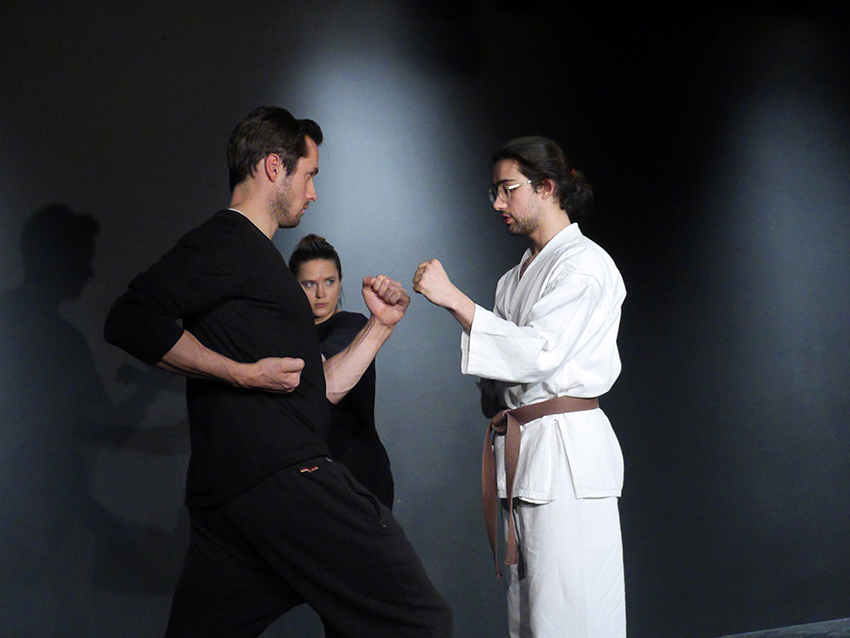 Links steht ein Mann in schwarzer Übungskleidung. Ihm gegenüber ist der Karatelehrer. Er zeigt ihm eine bestimme Haltung. Im Hintergrund schaut eine junge Frau, ebenfalls in schwarzer Übungskleidung interessiert zu.