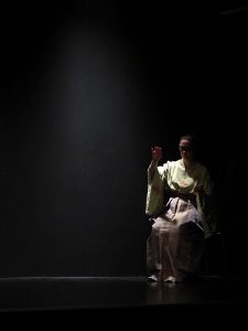 Die blinde Naoko sitzt in ihrem gelblichen und glitzernden Kimono rechts hinten auf einem Stuhl im Bühnenraum. Sie tastet vorsichtig in der Luft