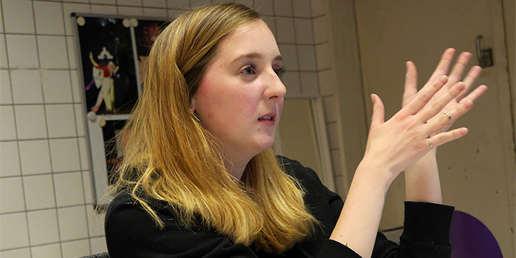 P1490415 web TW Kira im Gespräch. Eine junge Frau mit roten, langen Haaren sitzt an einem Tisch. Vor ihr steht ein Laptop und ein Glas Orangensaft. Sie ist konzentriert in einem Gespräch.