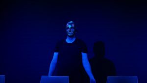 Ein Schauspieler steht hinter einer Stuhlreihe im blauen Bühnen licht. Er trägt eine goldene Maske