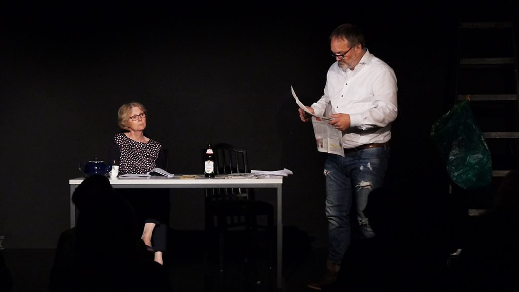 Links sitzt Lore (Sigird Abendroth) und schut in Richtung Harry (Marc Dauenhauer) er steht rechts vor dem Tisch und liest Zeitung. Die Bühne ist schwarz. Es gibt einen Tisch, zwei Stühle und einen gmütlichen Sessel auf der linken Seite.