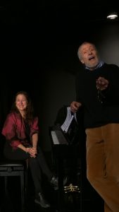 Sybille Karrasch am Klavier und Wolfgang Keuter steht vorne rechts im Bild. Beide lachen.