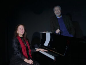 Wolfgang Keuter steht am Klavier, er trägt ein Tweedsakko und einen blauen Pulli. Sybille Karrasch sitzt am Klavier ist schwarz gekleidet und trägt einen roten Schal. Beide lächeln in die Kamera