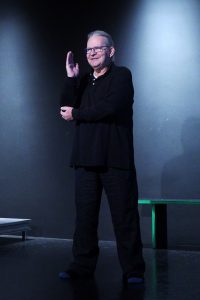 Ein Mann mit weißen Haaren, die zu einem Pferdeschwanz gebunden sind, zeigt eine asiatische Handgebärde. Er hat ein freundliches Gesicht.