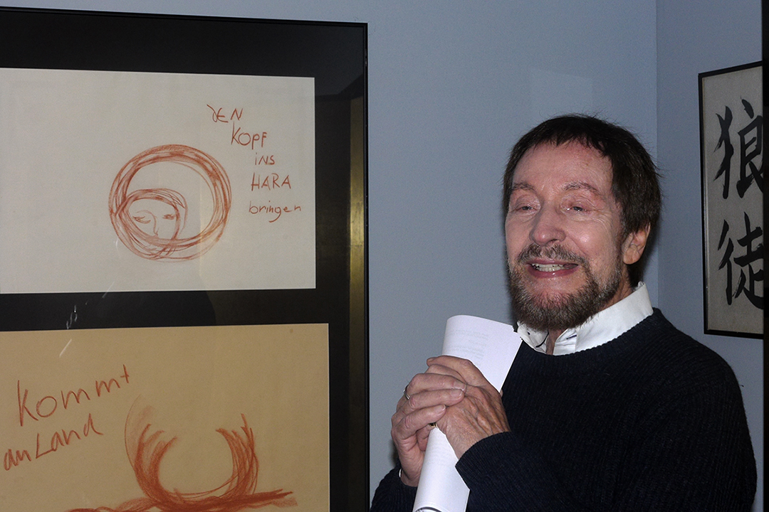 Wolfgang Keuter spricht über Slow Acting. Er trägt einen dunklen Pulli und ein weißes Hemd und steht im Studienraum. Links im Bild sind zwei Zeichnungen, auf der einen ist ein Gesicht zuerkennen, rechts davon steht „Den Kopf ins Hara bringen.“ zu sehen.