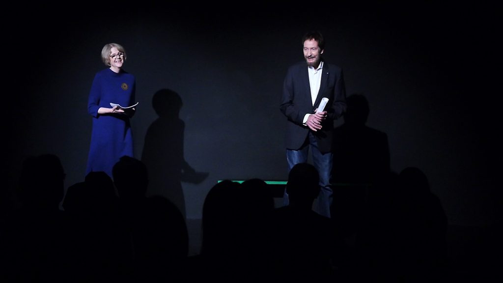 Sigrid Abendroth trägt ein blaues Kleid mit goldener Brosche, Wolfgang ein weißes Hemd und einen dunklen Blazer. Beide sind auf der Bühne und haben das Manuskript der dialogischen Lesung von Effi Briest in der Hand.