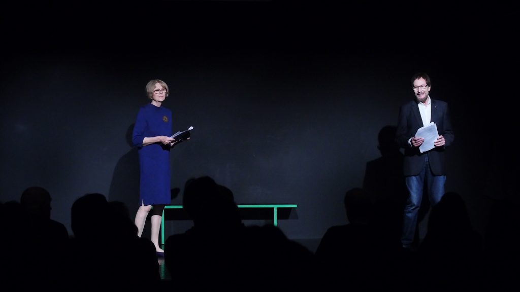 Sigrid Abendroth trägt ein blaues Kleid mit goldener Brosche, Wolfgang ein weißes Hemd und einen dunklen Blazer. Beide sind auf der Bühne und haben das Manuskript der dialogischen Lesung von Effi Briest in der Hand.