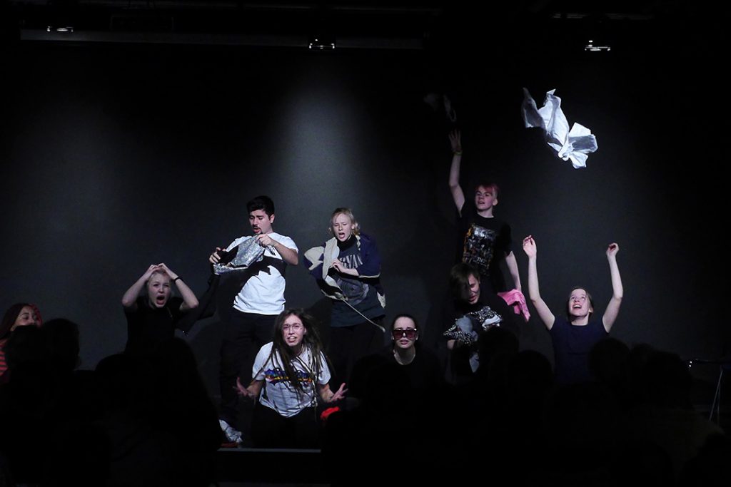 Neun Schüler zeigen eine Szene aus ihrem Programm Kabarettungsdienst KLIMAX. Das Thema ist der übermäßige Konsum von Kleidung. Viele Kleidungsstücke fliegen in hohem Bogen durch den Raum.