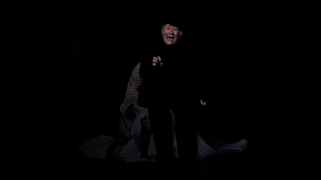 Im schwarzen Bühnenraum sitzt eine ältere Frau auf einem Stuhl und schreit mit einem entsetztem Gesicht.