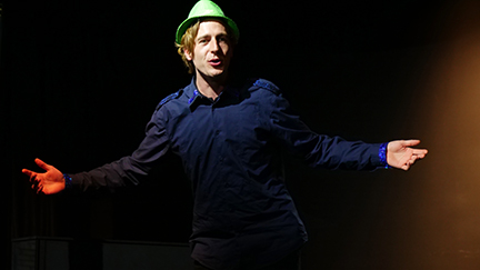 Szene aus dem Schauspieltraining. Ein Spieler auf der Bühne, er trägt einen grünen Glitzerhut und steht mit offenen Armen dem Publikum gegenüber