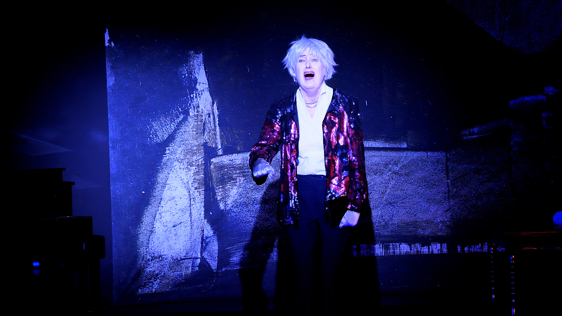 1111798_web-1024x768 Szene aus der Tor und der Tod. Eine Schauspielerin, die Mutter, steht mit glitzernder Jacke in der Mitte der Bühne. Sie hat einen starken gesichtsausdruck.
