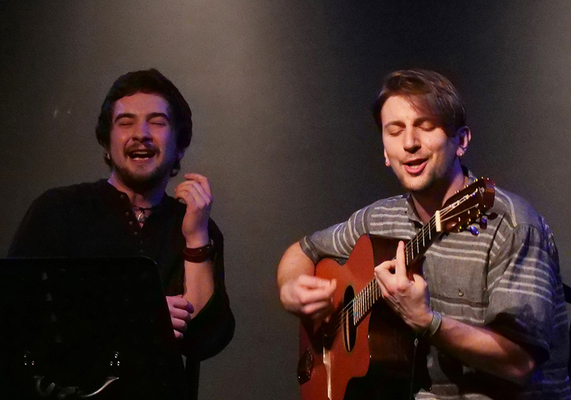 Zwei junge Männer sitzen auf der Bühne. Der linke singt emotional und der rechte spielt Gitarre.