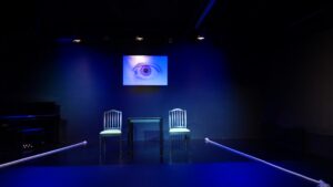 Schwarzer Bühnenraum. Im Hintergrund hängt ein großes Bild von einem geöffneten Auge in violett. In der Mitte steht ein Tisch mit zwei Stühlen. Links und rechts liegen jeweilws ein weißes auf dem Boden., welche den Bühneraum begrenzen.