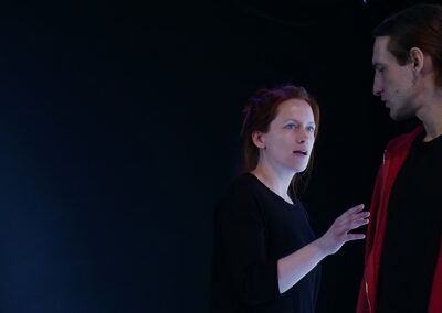 Eine Schauspielerin und ein Schauspieler in schwarzer Bühnenkleidung, bei Proben mit Text in der Hand.