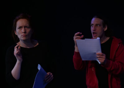 Eine Schauspielerin und ein Schauspieler in schwarzer Bühnenkleidung, er trägt noch eine rote Jacke, bei Proben mit Text in der Hand.