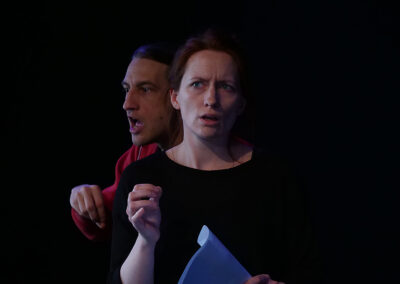 Eine Schauspielerin und ein Schauspieler in schwarzer Bühnenkleidung, er trägt noch eine rote Jacke, bei Proben mit Text in der Hand.
