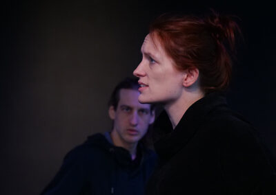 Eine Schauspielerin und ein Schauspieler in schwarzer Bühnenkleidung, bei Proben mit Text in der Hand.