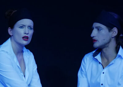 Ein Mann und eine Frau, beide tragen ein weißes Hemd oder eine weiße Bluse. Sie sind beide gleich hell geschminkt mit einem roten Mund und einem Eyelinerstrich unter den Augen. Sie proben eine Szene auf leerer, schwarzer Bühne.