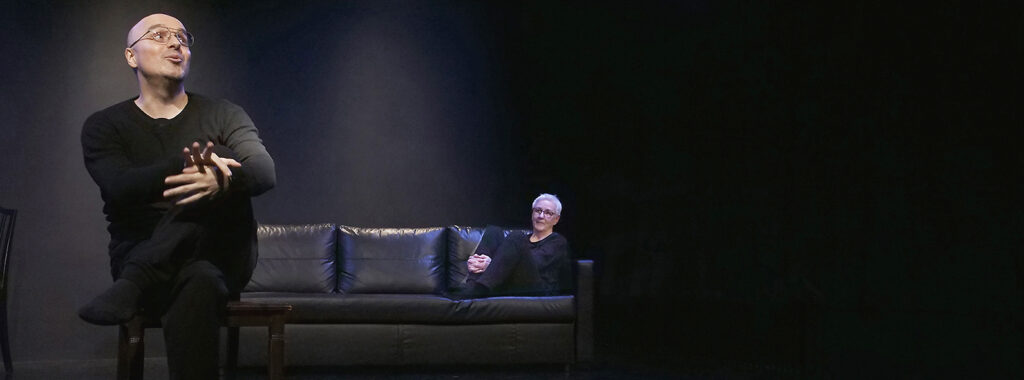 SChwarze Bühne mit schwarzem Ledersofa. Darauf sitzt hinten rechts eine Schauspielerin und beobachtet den Schauspieler, der vorne links auf einem Stuhl sitzt.