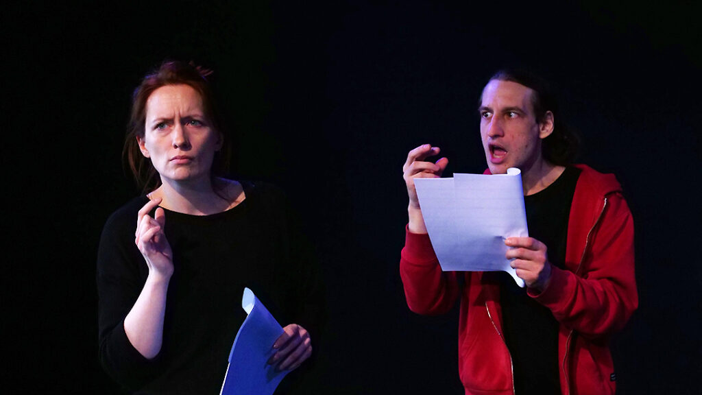 Zwei Personen beim Schauspieltraining auf der Bühne, beide in dunkler Kleidung, halten ein Skript in der Hand und probieren eine Szene unter Theaterlicht.