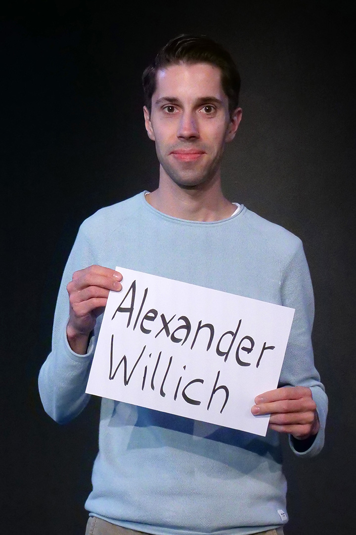 P1755793_web Alexander Willich, Beisitzer, er trägt ein hellblaues Shirt und lächelt in die Kamera. In den Händen hält er eine DIN A 4 Seite auf der sein Name steht.
