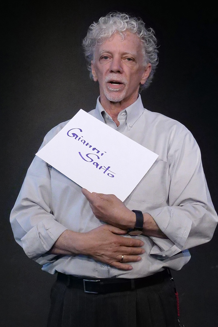 Gianni Sarto, Künsterlischer Leiter / QMB, er trägt ein graues Hemd und lächelt in die Kamera. In den Händen hält er eine DIN A 4 Seite auf der sein Name steht.