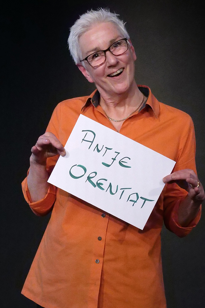 Antje Orentat, 1. Vorsitzende, sie trägt eine orange Bluse und lächelt in die Kamera. In den Händen hält sie eine DIN A 4 Seite auf der ihr Name steht.