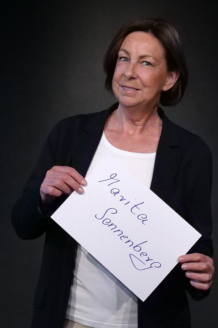 P1755830_web Marita sonnenberg, Beisitzerin, sie trägt ein weißes T-Shirt und eine dunkle Weste und lächelt in die Kamera. In den Händen hält sie eine DIN A 4 Seite auf der ihr Name steht.