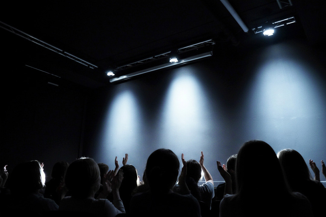 Blick von hinten auf die Silhouetten eines applaudierenden Publikums. Sie schauen auf eine dunkle, blau beleuchtete Theaterbühne, auf der zwei Stühle stehen und ein großes Auge auf einem Bildschirm gezeigt wird.