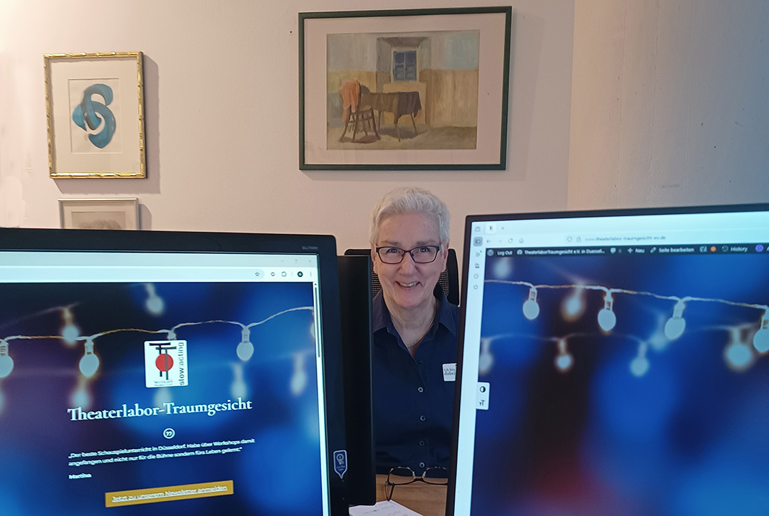 Antje Orentat sitzt lächelnd im Büro des TheaterLabor TraumGesicht zwischen zwei Monitoren an einem Schreibtisch, im Hintergrund hängen gerahmte Bilder.
